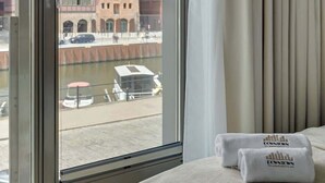 A hotel room with a large window, a bed, and a view of buildings and a canal. - Waterlane E2 | Spa, Pool & Gym | Riverside (Gdańsk)
