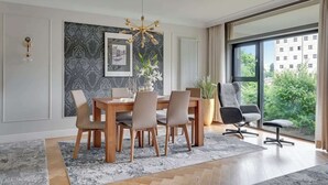 Un espace repas avec une table et des chaises en bois, un lustre et une vue sur la verdure grâce à de grandes fenêtres.