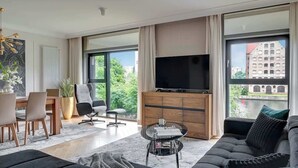 Un salon moderne doté d’un grand téléviseur, de sièges confortables, d’un coin repas et de grandes fenêtres offrant une vue sur les bâtiments et la verdure.