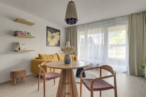 Ein modernes Wohnzimmer mit einem runden Holztisch, zwei Stühlen, einem gelben Sofa und einem großen Fenster mit transparenten Vorhängen.