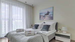 Ett modernt sovrum med en stor säng, ett sängbord, ett litet bord och ett fönster med tunna, genomskinliga gardiner.