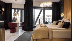 Moderni hotellihuone, jossa on suuri sänky, oleskelualue, jossa on punainen nojatuoli, kattokruunu ja suuret ikkunat, joista avautuu näkymä kaupunkiin.