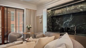 Moderni hotellihuone, jossa on sänky, pieni pyöreä pöytä maljakolla, sohva ja keittiöalue, jonka taustaseinä on marmoria.