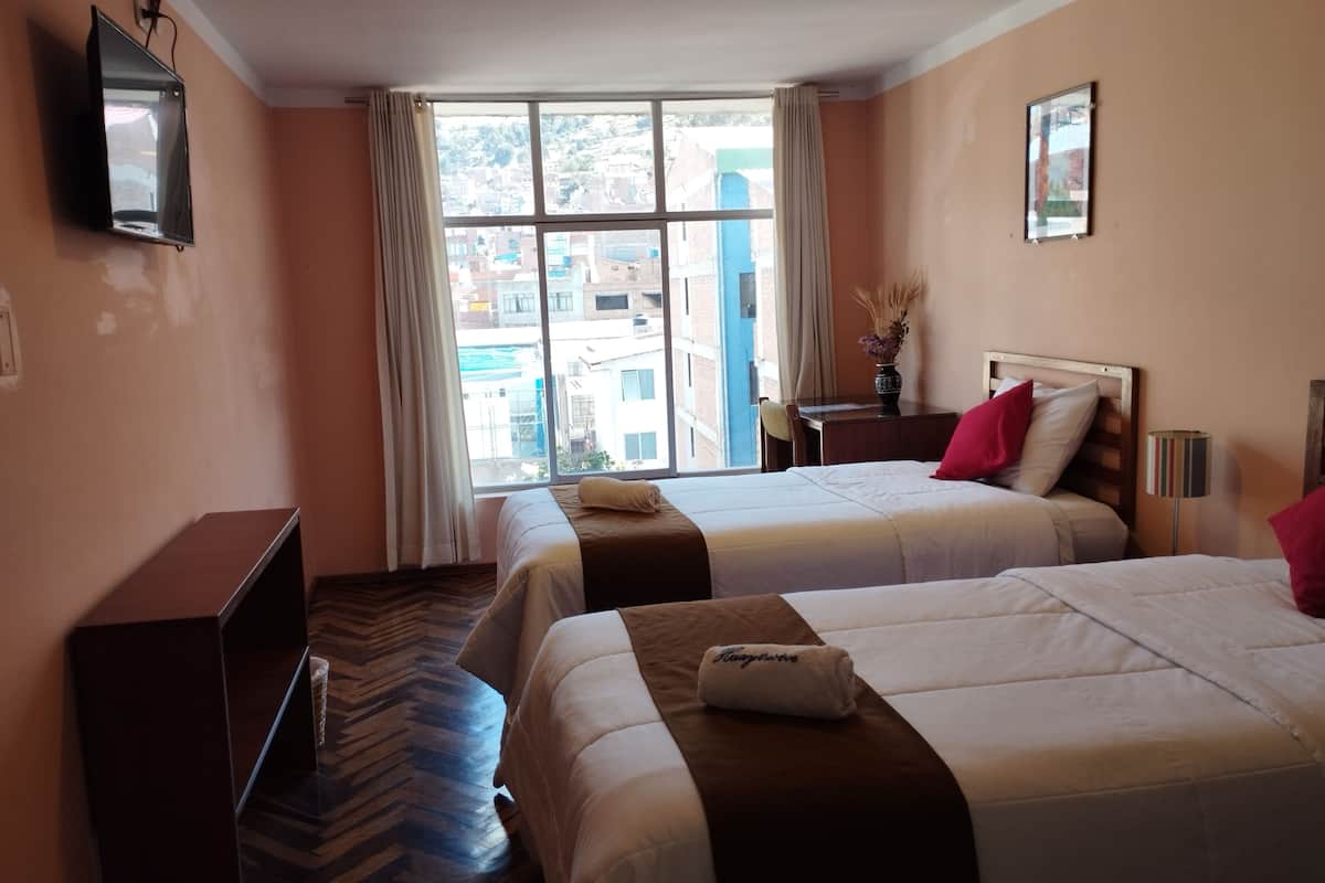 Ett hotellrum med två sängar, ett skrivbord, en TV och ett stort fönster med utsikt över byggnader.