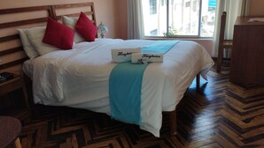 A hotel room with a bed, wooden headboard, red and white pillows, a blue throw blanket, and a view of buildings through the window. - Huaytusive (Puno)