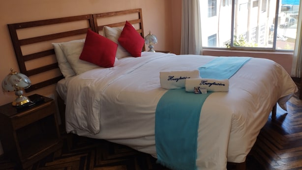 A neatly made bed with a wooden headboard, two red pillows, a white pillow, and a blue throw blanket. There are two bedside lamps on wooden nightstands. - Huaytusive (Puno)