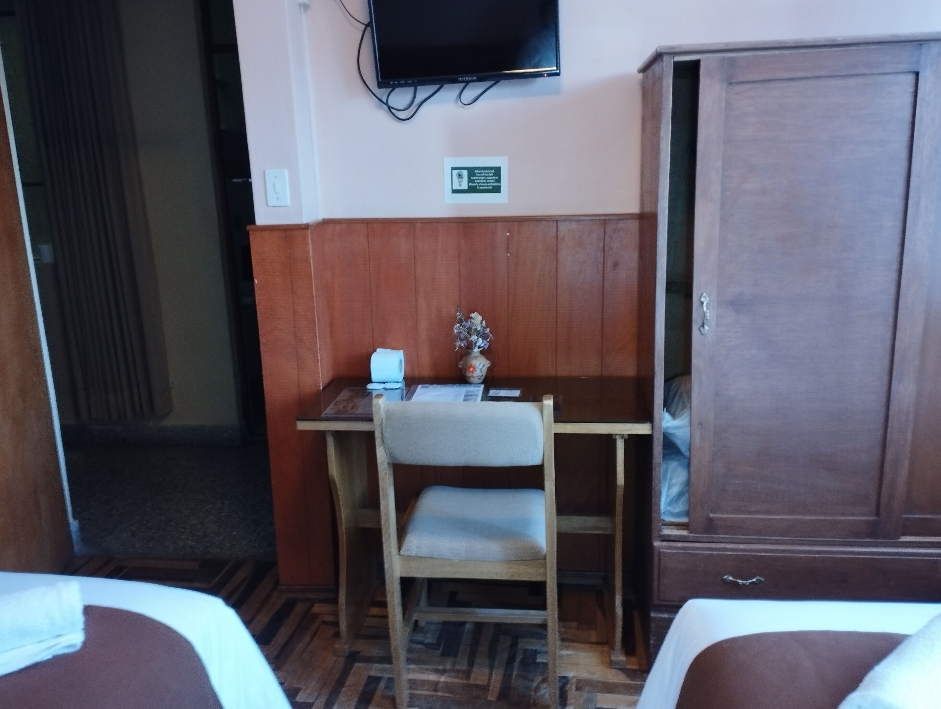 A hotel room with a wooden desk, a chair, a television mounted on the wall, and a wardrobe.