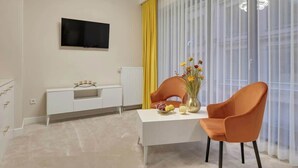 A modern living room with a flat-screen TV, a white console table, two orange chairs, a vase with flowers, and a large window with sheer curtains. - Granaria B106 | Hotel Standard Apartment (Gdańsk)