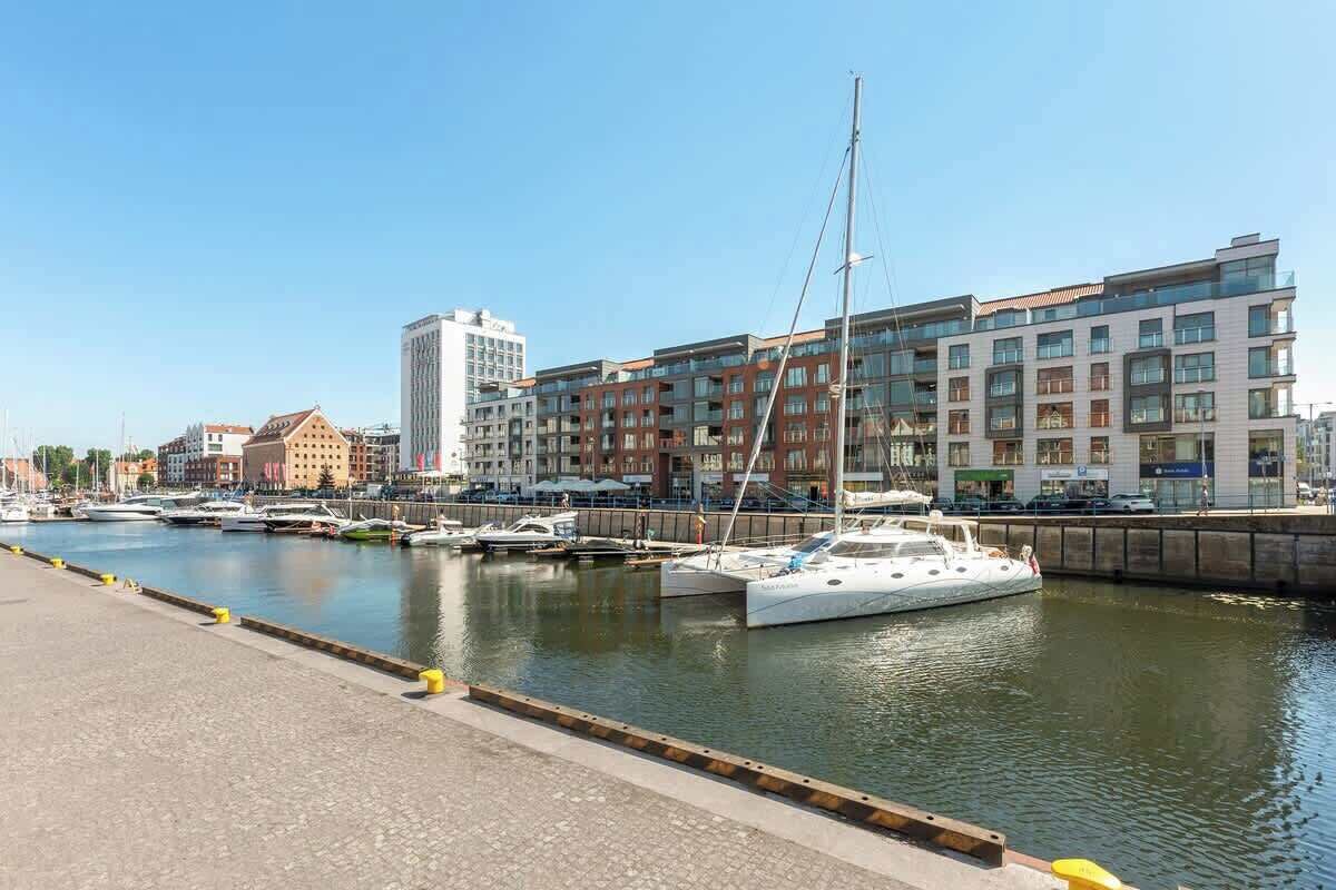 Eine Marina mit vertäuten Booten, eine Uferpromenade und moderne Gebäude im Hintergrund.