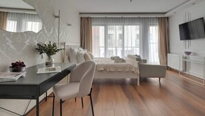 A modern hotel room with a large bed, a sitting area with a sofa and armchairs, a dining table with chairs, a large mirror, and a television mounted on the wall. - Waterlane F9B | Spa, Pool & Gym | Riverside (Gdańsk)