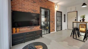 A modern living room with a brick wall, a flat-screen TV, a dining area with stools, and a bar counter. - Granaria A212 | Best Location (Gdańsk)
