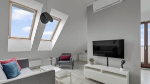 Una sala de estar moderna con una televisión de pantalla plana, un sofá, una mesa de centro y una silla. Hay tragaluces y un equipo de aire acondicionado en la pared.