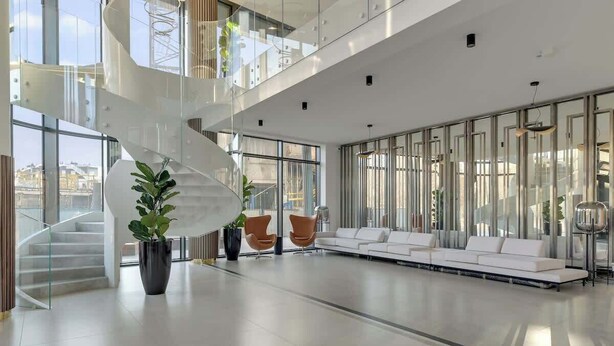 Een ruime lobby met een centrale spiraaltrap, een witte bank, oranje fauteuils en grote ramen.