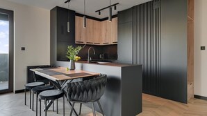Een moderne keuken met een centraal eiland, zwart en houten kastjes, hanglampen, en een eethoek met een tafel en stoelen.