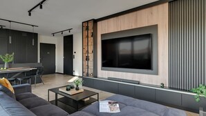 Een moderne woonkamer met een grote flatscreen televisie aan een houten wand, een donkergrijze bank, een salontafel en een eethoek met een zwarte stoel.