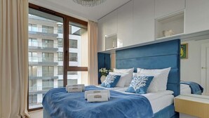 A modern hotel room with a large bed, a bedside table, a nightstand, a window with curtains, and a view of a building outside.