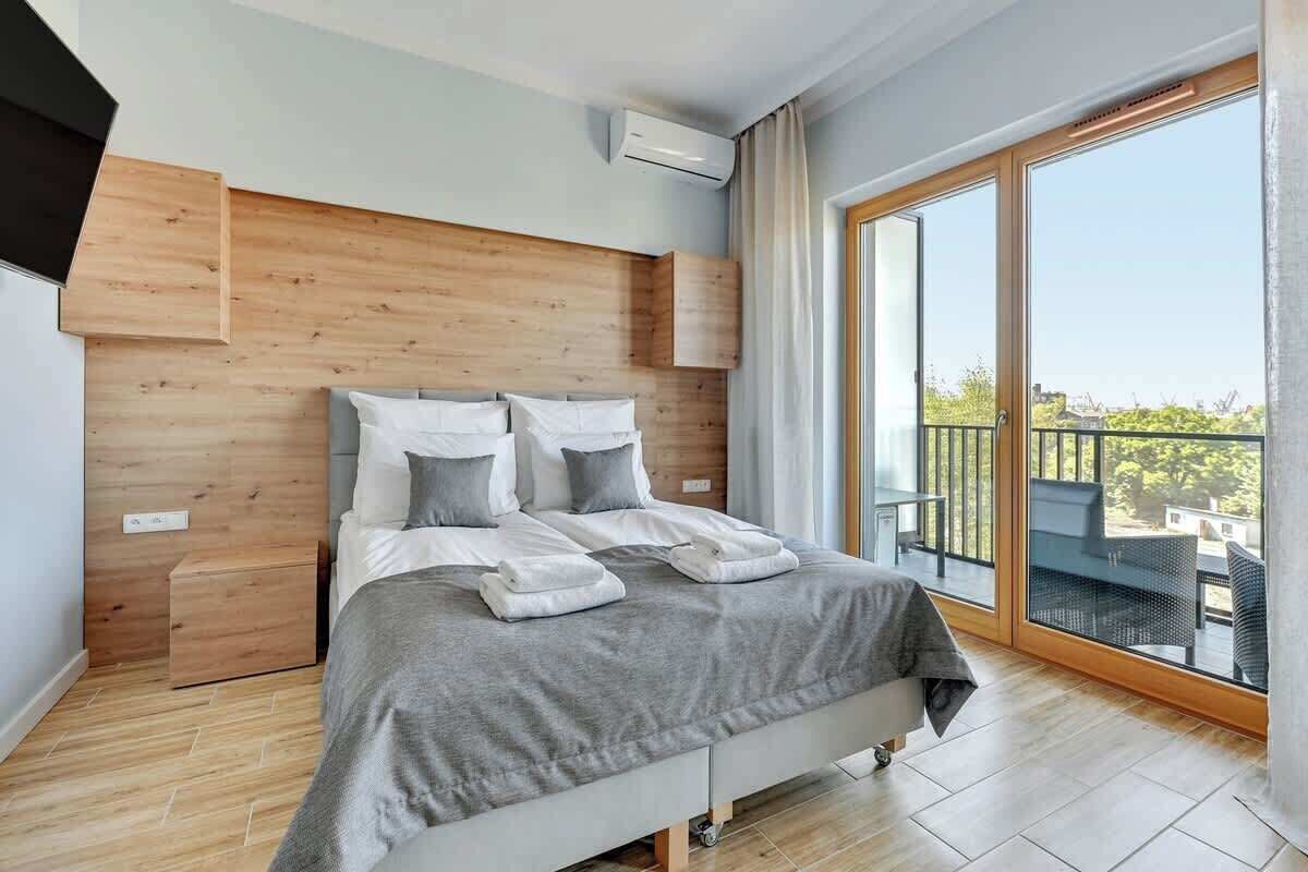 Een moderne slaapkamer met een houten hoofdbord, een groot bed met wit en grijs beddengoed, een klein nachtkastje en een balkon met uitzicht.