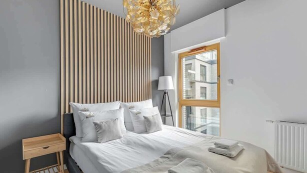 Een moderne slaapkamer met een bed, nachtkastje, lamp en uitzicht op gebouwen door een raam.
