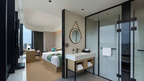 Une chambre d’hôtel moderne équipée d’un lit, d’une table de chevet, d’un miroir et d’une douche séparée par des parois en verre.