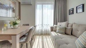 En moderne stue med sofa, spisebord med stole og et stort vindue med gardiner.