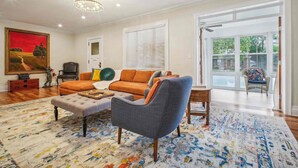 A spacious living room with a large sofa, a grey armchair, and a coffee table. There is a painting on the wall and a window with blinds. - Spacious 4-bedroom Single Family home in Historic  Weequahic Newark NJ (Newark)