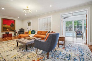 A spacious living room with a large sofa, a grey armchair, and a coffee table. There is a painting on the wall and a window with blinds. - Spacious 4-bedroom Single Family home in Historic  Weequahic Newark NJ (Newark)