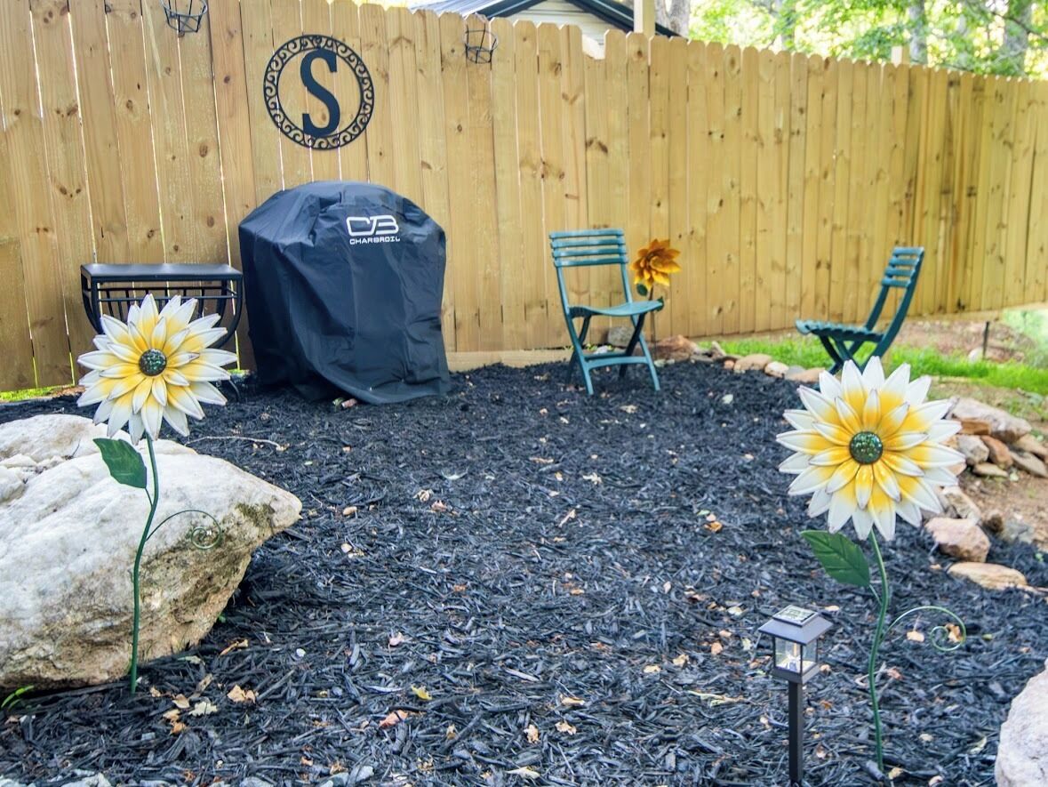 A backyard with a wooden fence, a barbecue grill, two green chairs, and two artificial sunflowers.
