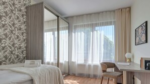 Ein Hotelzimmer mit einem Bett, einem Schreibtisch, einem Stuhl, einem großen Fenster mit durchsichtigen Vorhängen und einer Wand mit einem Blattmuster-Tapezierpapier.