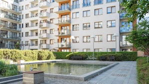 Ein mehrgeschossiges Wohnhaus mit Balkonen, ein gepflasterter Innenhof mit einem kleinen Teich und einer Bank.
