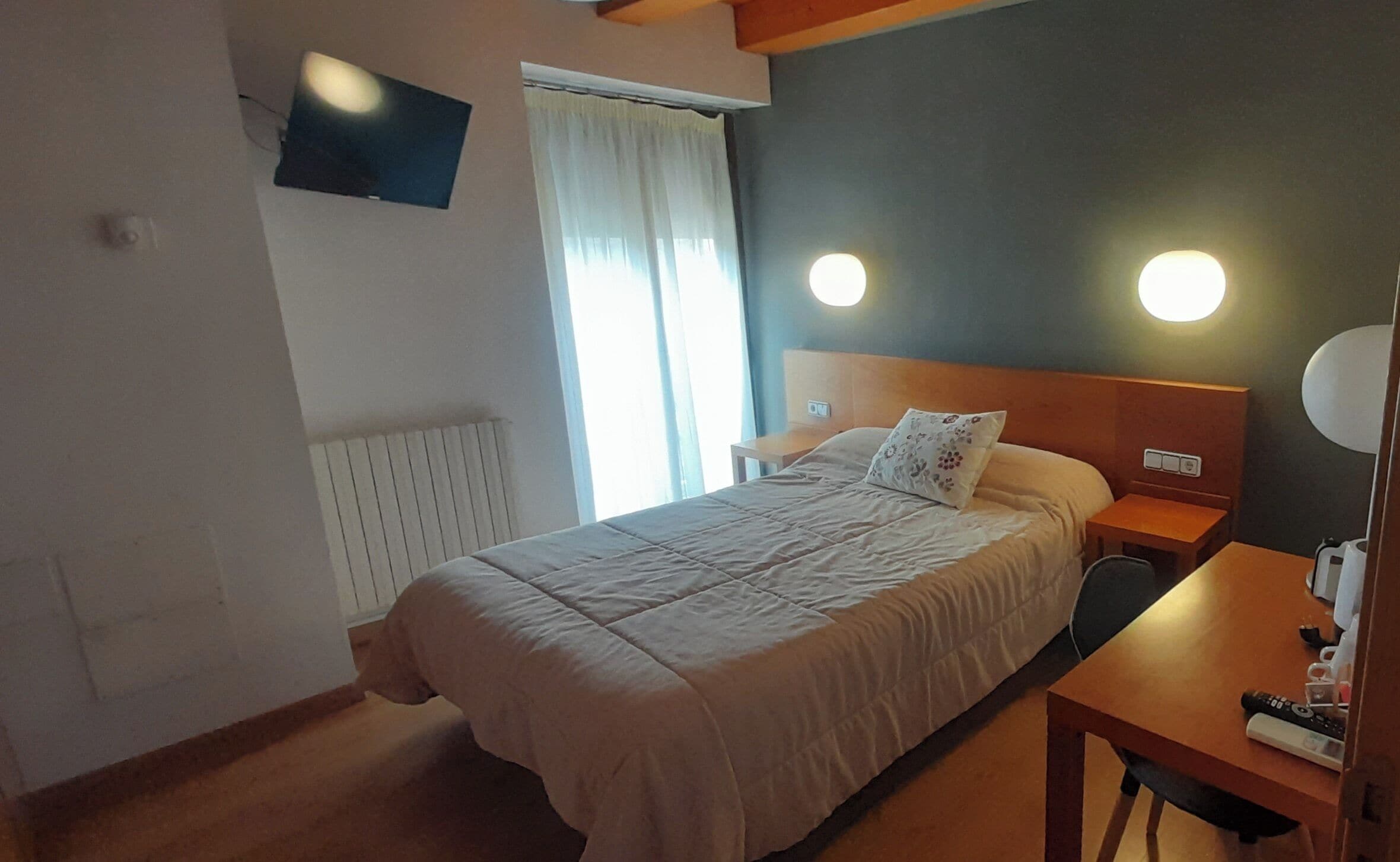 A hotel room with a bed, bedside tables, a TV mounted on the wall, and a window with curtains.