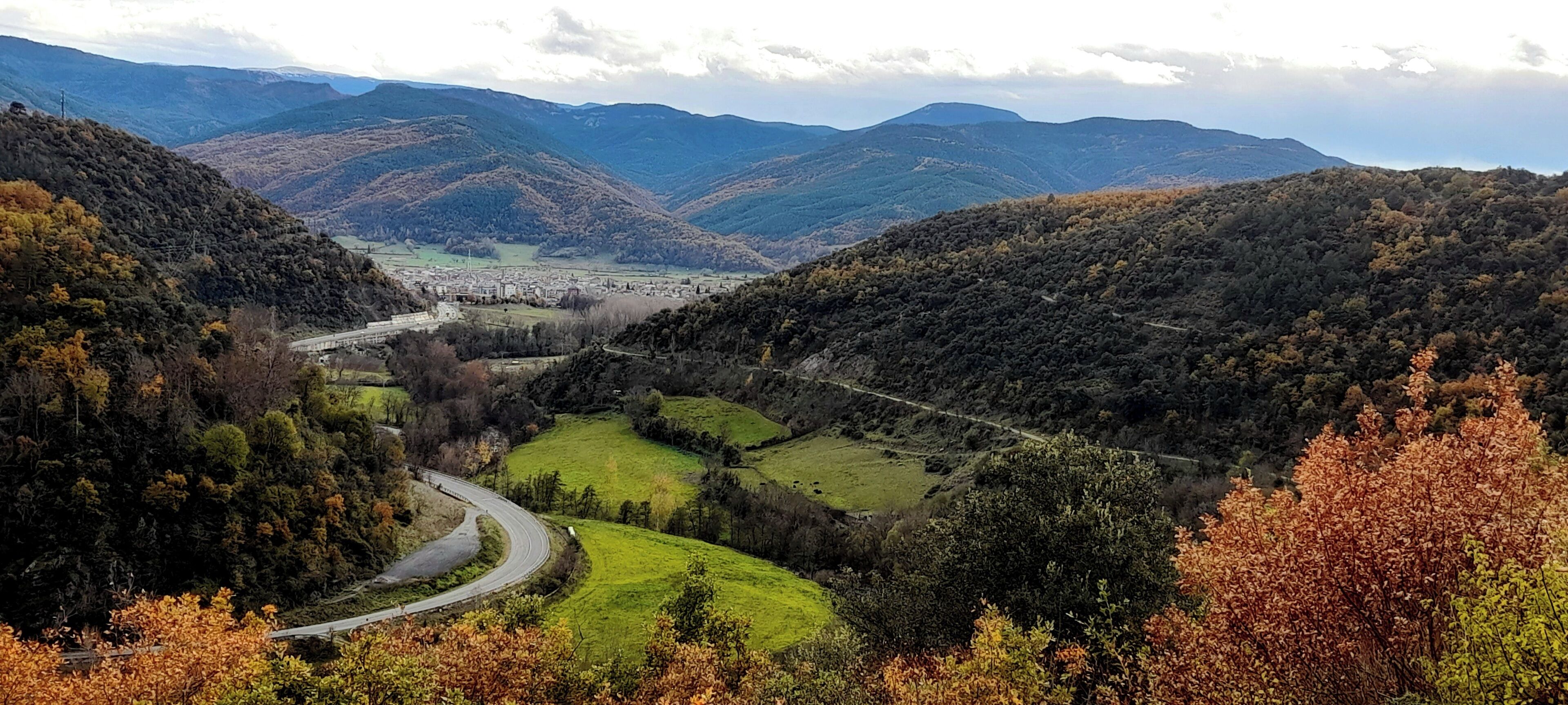 Un paisaje montañoso con un camino serpenteante, campos verdes y colinas distantes.