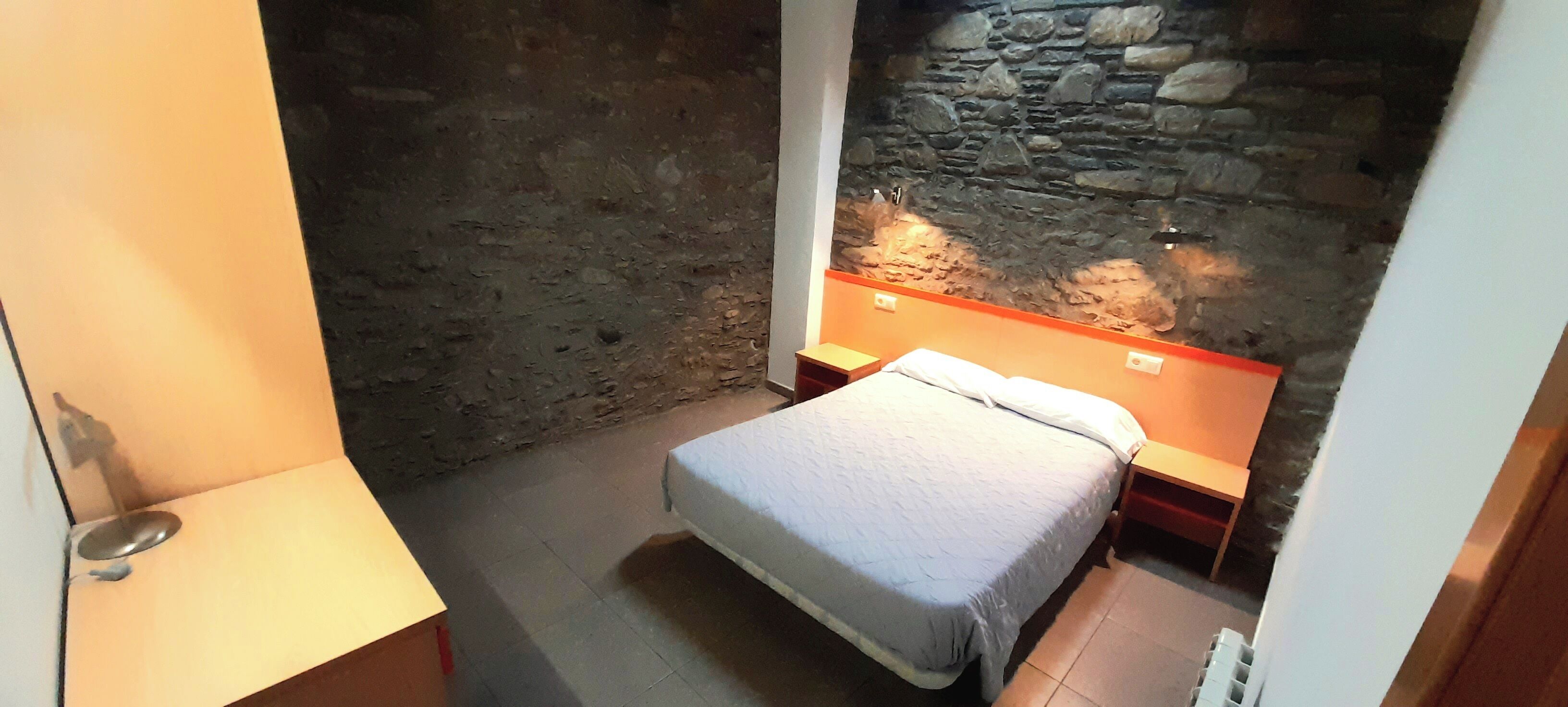 Una stanza piccola e stretta con una parete in pietra, un letto singolo con materasso bianco, un comodino e una lampada da notte.