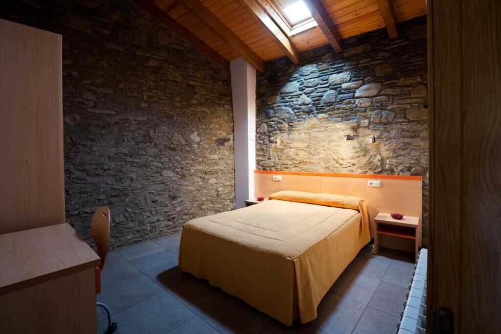 A bedroom with a stone wall, wooden ceiling beams, a bed with a beige bedspread, a nightstand, and a desk.