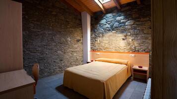 A bedroom with a stone wall, wooden ceiling beams, a bed with a beige bedspread, a nightstand, and a desk.