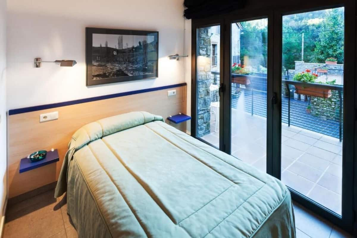 A bedroom with a bed, a nightstand, a wall-mounted picture, and a sliding glass door leading to an outdoor area with plants and a bench.
