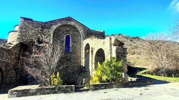 Una estructura antigua de piedra con ventanas arqueadas y un techo empinado, rodeada de árboles y un cielo despejado.