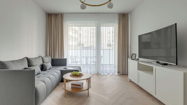 Ett modernt vardagsrum med en soffa, ett soffbord, en tv som står på ett skåp och ett stort fönster med gardiner.