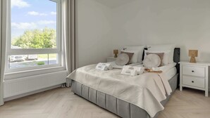 Ett sovrum med ett stort fönster, en säng med ett vitt och grått täcke, ett sängbord med en lampa och ett element under fönstret.