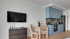 Une chambre moderne équipée d’un téléviseur à écran plat, d’une table à manger en bois avec deux chaises, d’une commode sombre et d’un coin cuisine avec des placards bleus et une cuisinière.