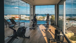 Ein geräumiges Zimmer mit großen Fenstern, die einen Pool und die Stadtlandschaft überblicken, ausgestattet mit Fitnessgeräten.