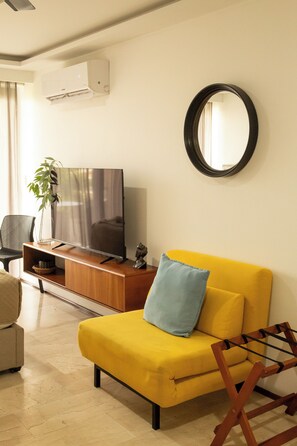 A living room with a yellow sofa, a TV on a wooden stand, a round mirror on the wall, and a plant. - Rooftop Pool Studio · Steps from 5th Ave & Beach (Playa del Carmen)