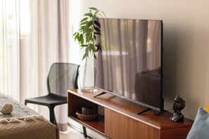 A living room with a flat-screen TV on a wooden stand, a black chair, a green plant, and a decorative item on the stand. - Rooftop Pool Studio · Steps from 5th Ave & Beach (Playa del Carmen)