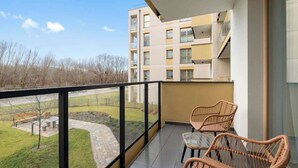 Een balkon met rieten stoelen, uitzicht op een park, en een gebouw op de achtergrond.