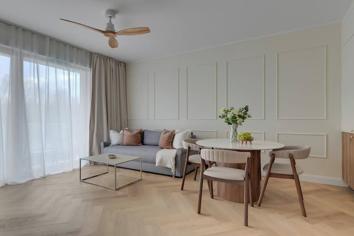 A modern living room with a sofa, a dining table with chairs, a coffee table, a ceiling fan, and a window with sheer curtains.