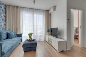 Un salon moderne avec un canapé, un pouf, une télévision et une petite table sur laquelle est posée une plante.