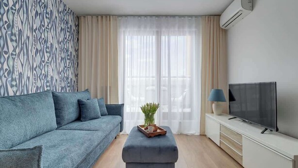 Un salon moderne avec un canapé, un pouf, une télévision et des rideaux.
