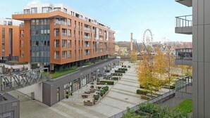Ein modernes Mehrstöckiges Gebäude, ein Innenhof mit Sitzgelegenheiten und ein Riesenrad im Hintergrund.