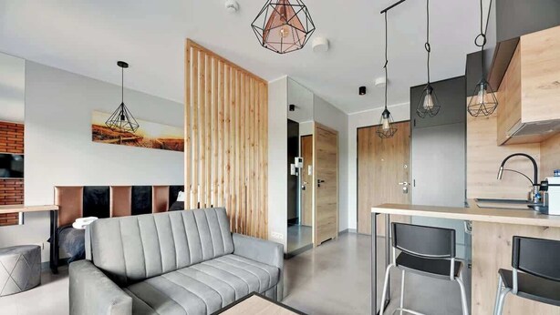 Una sala de estar moderna con un sofá gris, separador de listones de madera y una zona de cocina con taburetes de bar.