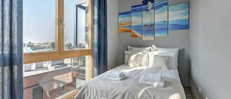 A bedroom with a large window, a bed with white linens, and three paintings on the wall.