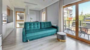 Un soggiorno moderno con un divano color petrolio, un pouf rotondo, un’area pranzo con sedute da esterno e un’ampia porta scorrevole in vetro che dà sull’esterno.